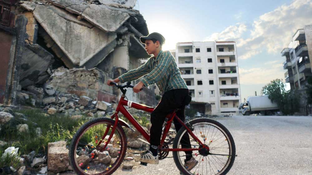 boy riding bicycle next to bombed building