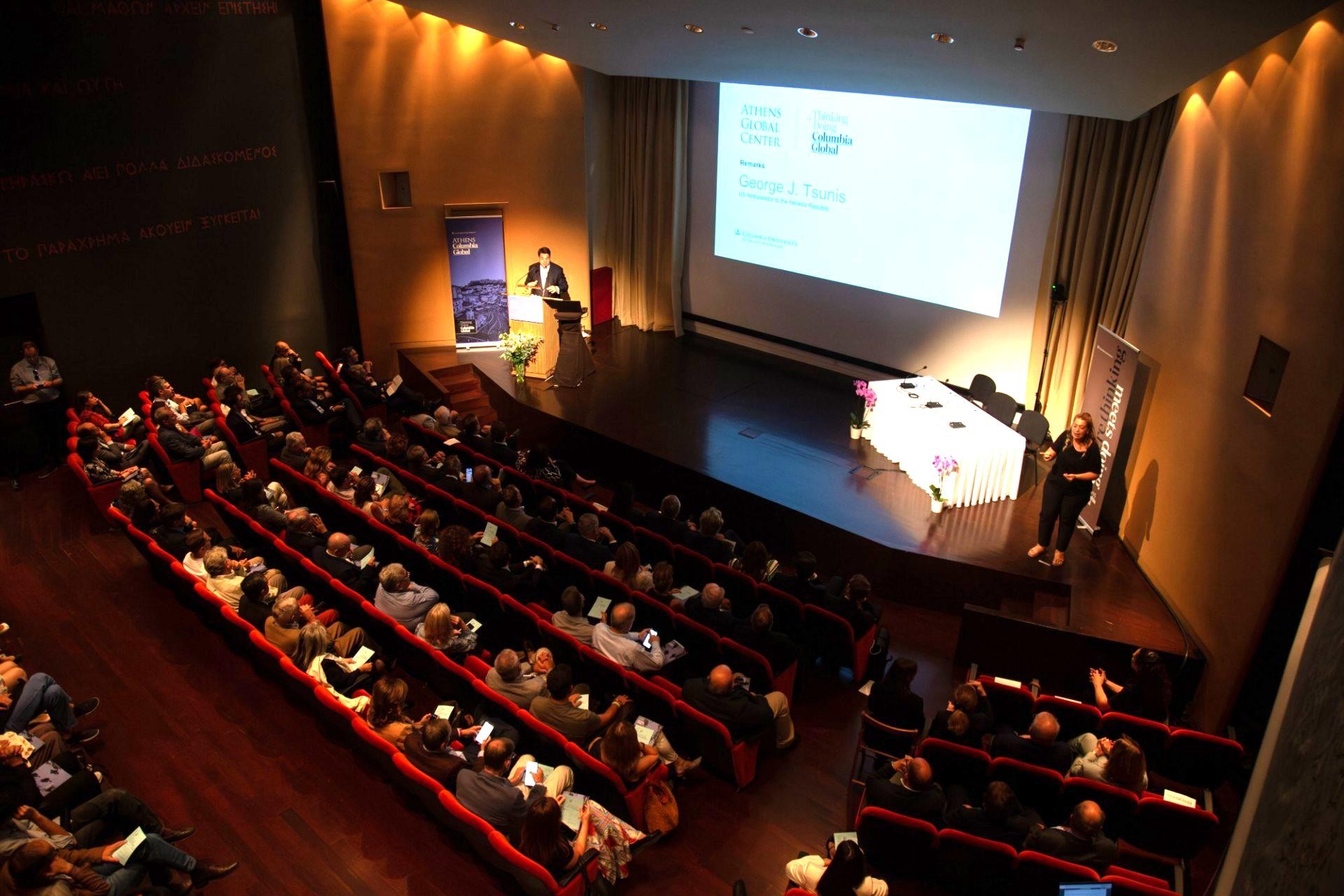 Athens auditorium Crowd in auditorium watches speaker in front of a presentation screen.