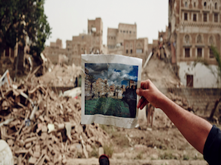 person standing in front of building destruction holding a photo of the building before it was destroyed