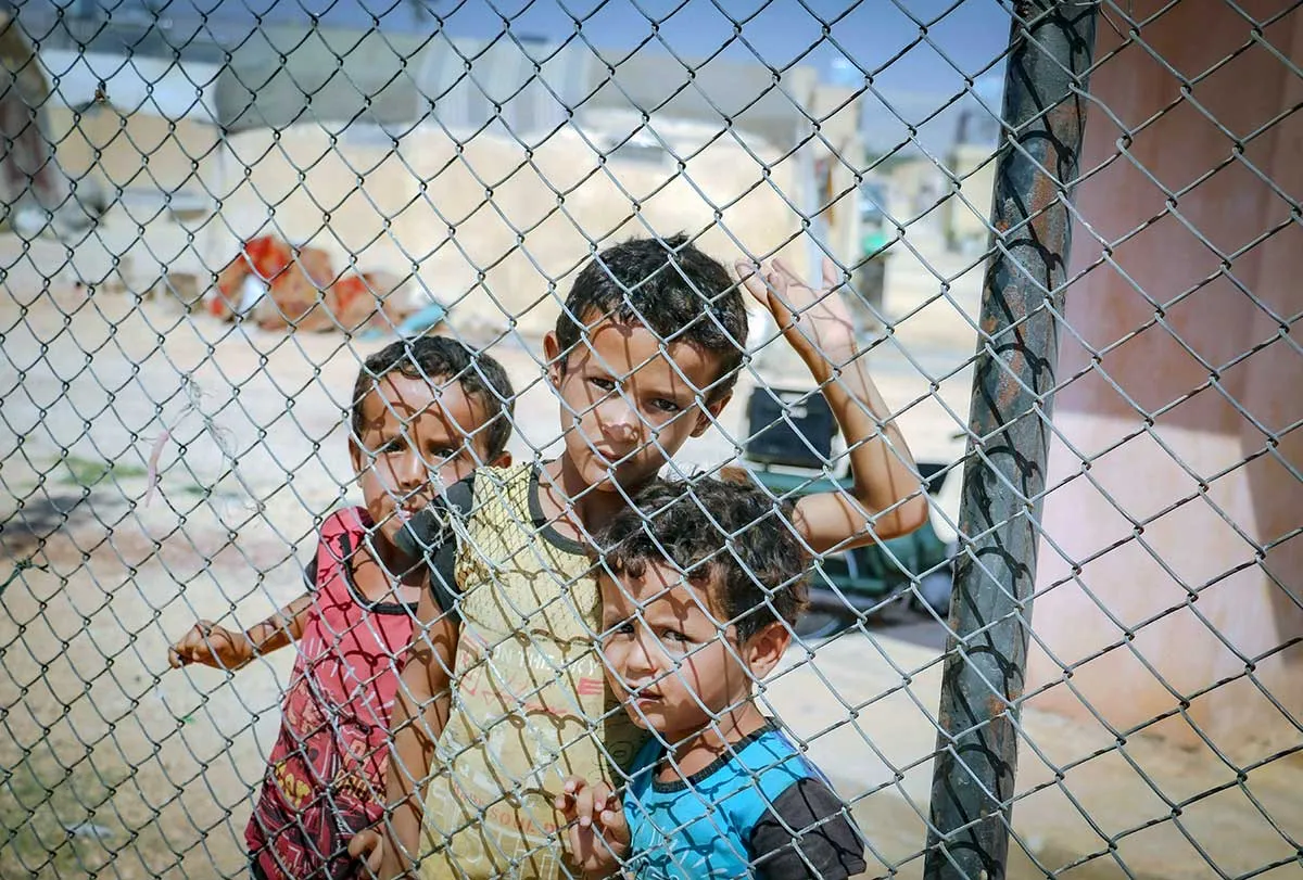 three children behind a fence at a refugee camp